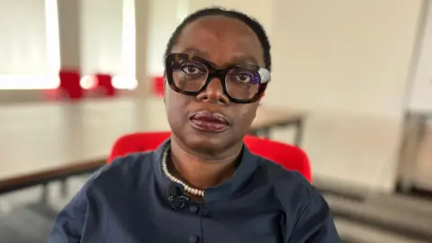 Martin Giles/BBC A woman sitting in a red chair within a room looks at the camera. The background behind her is blurred. She has dark, short hair, and is wearing large glasses along with a navy blue blouse and pearl-style necklace.