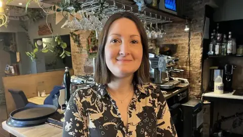 A woman in a patterned shirt stands smiling behind a cosy bar counter, with hanging glasses, bottles and a coffee machine in the background.