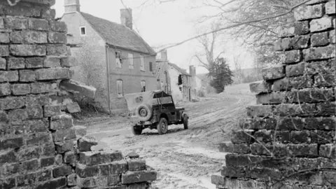 Hulton Archive/Getty Images An archive black and white picture showing a military-style car with a canvas roof driving through the abandoned village in the 1940s. There is a brick wall in the foreground which has collapsed and a house in the background has boarded up windows and damage to the walls.