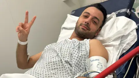 Pouria Zeraati lies in a hospital bed, wearing a hospital gown and wrist band, looking at the camera with two fingers raised in a peace sign