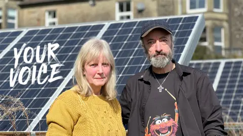 BBC Peter and Penney stand in front of solar panels in their garden in front of a stone house. She has should lenght blonde hair and a yellow sweather, while he has grey beard and dark cap with a black shirt openand showing a t shirt wi the muppet character animal drumming
