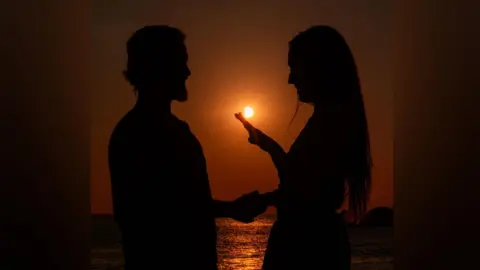 Ben Fiore The silhouette of a bearded man is on the left of the shot . He is holding the hand of a woman on the right of the shot. The sun is in the background behind them, with the sea in the distance.