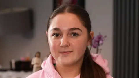 Erin Griffiths has long dark hair in a pony tail and dark eyes. She is wearing a pink hoodie and is looking directly at the camera