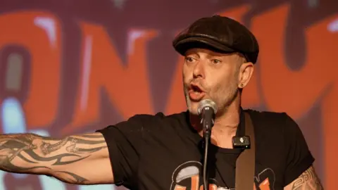 A man with tattoos and a black tshirt and cap behind a microphone with one arm outstretched and an orange painting on the wall behind him