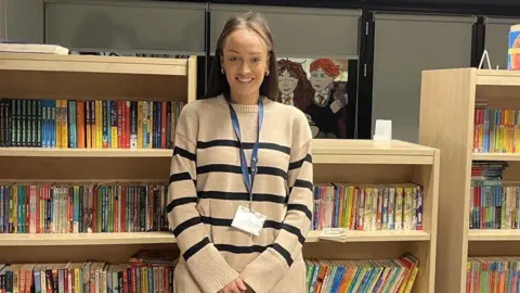 Amber Stapleton, looking at the camera, standing in front of a full bookcase in a school classroom