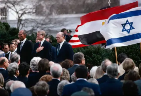 Getty Images Presidenti egjiptian Anwar El-Sadat, Presidenti i SHBA Jimmy Carter dhe kryeministri izraelit Menachem Fillojnë teksa qëndrojnë për himnet e tyre kombëtare përkatëse në lëndinë veriore të Shtëpisë së Bardhë, Uashington DC, 26 mars 1979. Pas Marrëveshjes së Camp David , ata ishin atje për të nënshkruar Traktatin e Paqes Egjipt-Izrael.