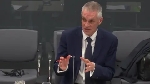 Senedd Cymru BBC director general Tim Davie addressing the Senedd culture committee. He is wearing a dark blue suit and tie and a white shirt and gesturing with his hands as he explains a point.