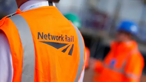 PA Media The back of someone wearing a Network Rail orange sleeveless top. There is a blurred image in the background of another person wearing a long sleeved orange coat and a blue hard hat.