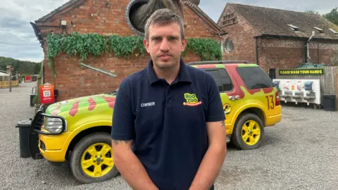 A man with grey/brown hair wearing a navy t-shirt that says "hoo zoo owner". There is a bright vehicle behind him that is yellow and green with brown animal patterns on it. On a building behind him is a large dinosaur model bursting out of the wall