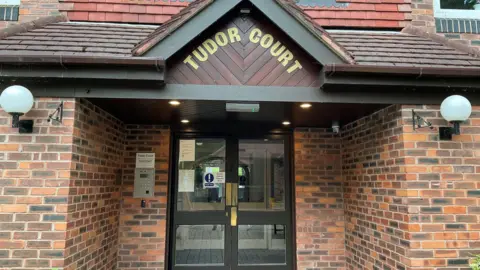 The outside of Tudor Court. It shows a brick entrance, glass doors and has the sign Tudor Court in gold writing at the top.