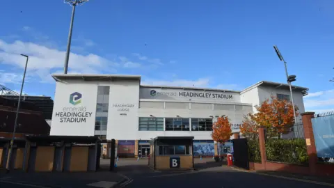 The exterior of Headingley Stadium is shown with its large white panels, glass windows and tall floodlights rising above the building. A gated entrance and autumn‑coloured trees sit in the foreground, making the stadium look both modern and familiar as a major Leeds sports venue.