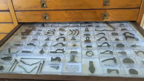 Nettie Edmondson A draw full of items found on searches, in a drawer. They are all in plastic boxes with writing by them. They are in rows and include different items. 