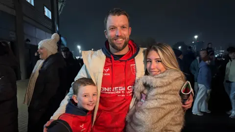 A tall man with a dark beard stands with two children either side him - a boy with short brown hair and a teenage girl with long blonde hair and a fur coat. The man and boy wear Hull KR kit.