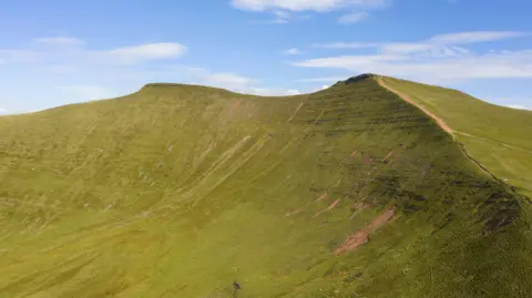 Getty Images The green hills and slopes of Pen y Fan seen on a nice day. The sky is blue above the hills.
