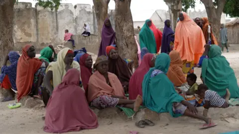 A group of more than a dozen women, many wearing brightly coloured hijabs, sit around a tree on dusty ground