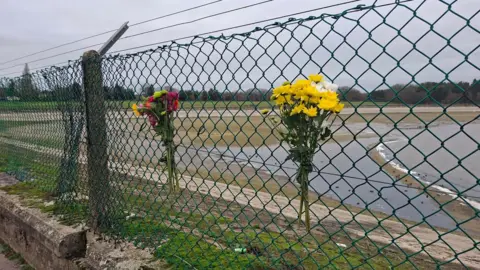 Henry Godfrey-Evans/BBC Two floral tributes left on a fence outside of a reservoir. The flowers are a mixture of colours.