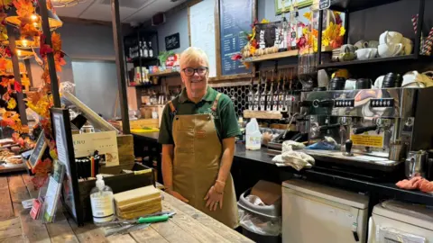 Liz Roger, die kort blond haar heeft en een bril met een zwart montuur draagt, een groen T-shirt met col en een beige schort, lacht naar de camera van achter de toonbank in Coffee Carriage, met een zilveren koffiezetapparaat, kopjes en flessen siroop zichtbaar op de achtergrond. 