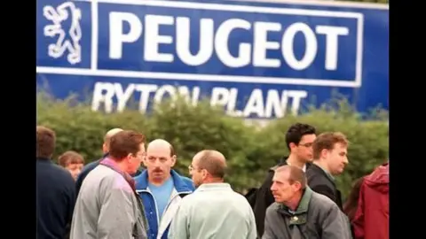 A group of men and one women stand outside in coats in front of a large blue sign saying Peugeot Ryton Plant.