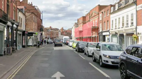 A street with cars parked down one side and a number of shop fronts on either side, with one building painted white and another painted pink