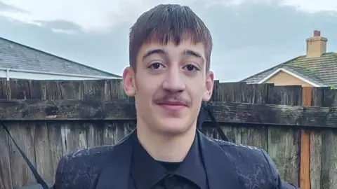 Taylor Charlton is a young man standing outdoors in front of a wooden fence. He is wearing a dark buttoned shirt and a dark patterned jacket. He has short dark hair and a light moustache. Behind the fence are the roofs of nearby houses and a cloudy sky.