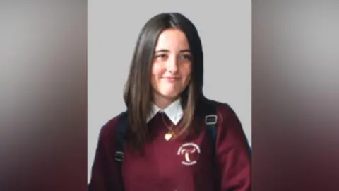 An image of a girl, wearing a burgundy school uniform and white shirt. She has long brown hair. There is a grey wall behind her. 