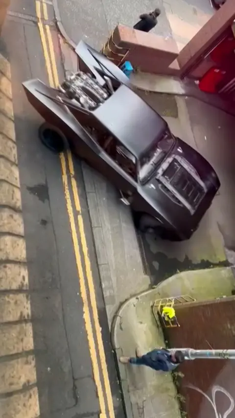 Overhead view of a large, angular black Batmobile parked on a narrow Glasgow street beside double yellow lines. Its engine is exposed at the rear, and several people stand nearby.