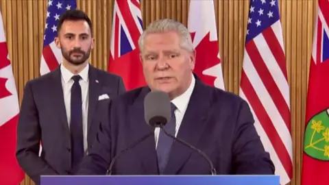 Doug Ford wears a blue suit and white shirt with a blue checked tie while speaking into a microphone.