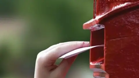 BBC A hand placing a letter into a round, red post box against a blurred green background.