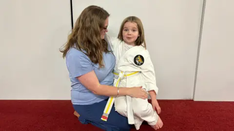 A woman kneeling on a red-carpeted floor, holding a child dressed in a white martial arts uniform with a yellow belt and a patch on the chest, against a plain light-coloured wall.