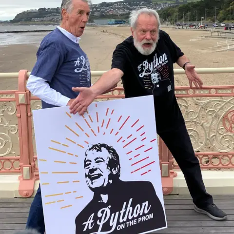 Sir Michael Palin and Terry Gilliam strike silly poses on Colwyn Bay pier while holding a poster featuring Terry Jones and advertising the Python on the Prom campaign
