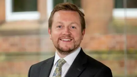 Dr Ian Collen A man smiling at the camera wearing a suit and tie. The jacket is black and a white shirt with a floral tie. He has ginger hair and a beard.