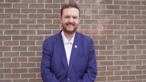 Alex McIntyre standing in front of a brick wall with a close beard and moustache, wearing a blue suit with a gold pin over a white shirt. 