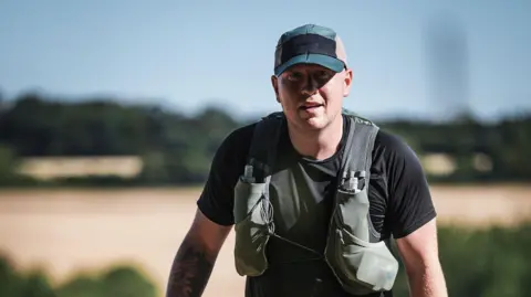 A man wearing a black t-shirt, a green-grey running vest with water bottles in pouches on either side of his chest and a blue hat is pictured on a hot day.