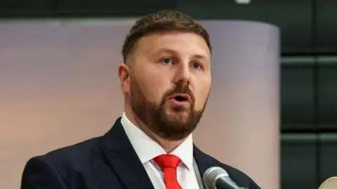 Reuters Chris Webb speaks into a microphone at an event. He wears a dark suit and red tie.