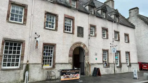 Dunbar's Hospital is a three storey building with white harled walls and small, lattice windows framed by exposed bricks. There is an arched doorway with an open metal gate.