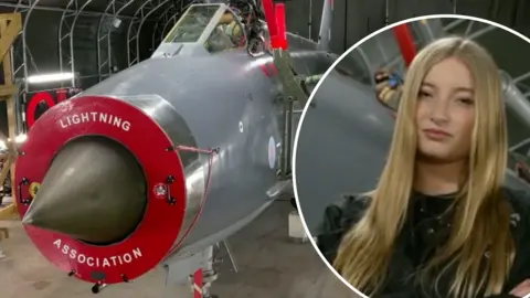 Girl with blonde hair with arms crossed wearing a black flight suit is captured next to a restored grey and red fighter jet