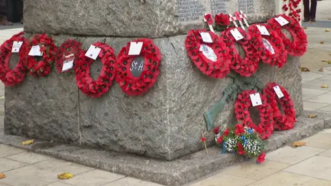 Poppy wreaths at the across the bottom of a war memorial in Cullompton. The poppy wreaths are red and have notes attached. 