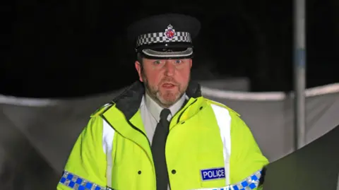 PA Media Ch Supt Neil Blackwood, wearing a yellow hi-vis Greater Manchester Police jacket, updates reporters at a media conference held in front of a police cordon.