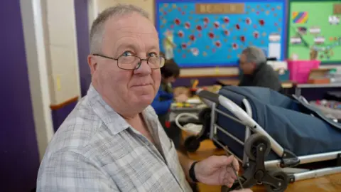 BBC A man with glasses and grey hair and wearing a grey check shirt holds a metal rod as he tries to fix a silver and blue trolley.