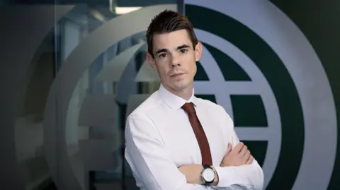 IWF Dan Sexton standing in front of a company logo. He is looking at the camera and is wearing a white shirt and a burgundy tie. He has short, dark hair