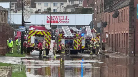 Três bombeiros e vários bombeiros trabalham para reduzir as enchentes em Coalisland, condado de Tyrone. A rua fora das fileiras de propriedades de tijolos vermelhos está inundada e há uma grande loja ao fundo.