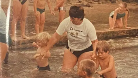 The Guernsey Swimming Club A yellow faded photo showing a woman with dark short hair in a white T-shirt, holding hands with two boys in a shallow pool.