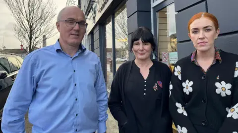 Three members of staff rom one of Kilrea's pharmacies - on the left is a bald man with glasses wearing a light blue shirt with black buttons, in the centre a woman with short black hair wearing a black blouse with a black cardigan over it and on the right a woman with red hair pulled back behind her head wearing a black cardigan with a daisy pattern on it.