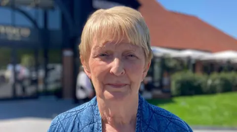 A woman with blonde hair and wearing a blue jacket looks into the camera. She is aged approximately 70. 