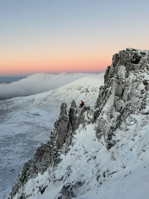 Eloise Simpson Portrait image of a pale blue sky, fading into orange and pink with snow covered mountains below. In the distance, a person is visible sitting on the peak of the mountain.