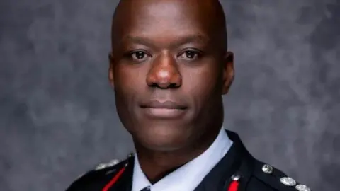 A man with a shaved head is looking at the camera. He is wearing a senior fire officer's uniform.