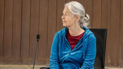 A woman with greying hair sits in front of a microphone, wearing a blue zip-up sweatshirt and red undershirt