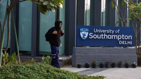 Reuters A man wearing a brown hoodie and blue trousers passes by the entrance to the campus. On his side is a board which reads University of Southampton Delhi. There are trees and shrubs on the side and a glass building in the backdrop. 