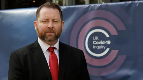 Eddie Lynch standing looking at the camera with a serious expression on his face. He is wearing a suit and tie. Behind him is a banner with the logo of the UK Covid-19 inquiry