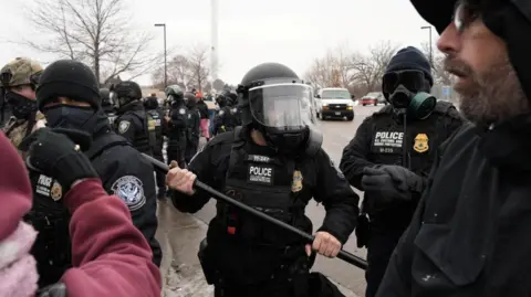a Customs and Border Patrol agent holding up a baton as more agents gather behind him